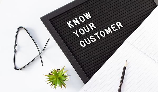Black letter board with ‘Know your customer’ in white letters, surrounded by black glasses, a green cactus, an open white notebook with a black pencil on it